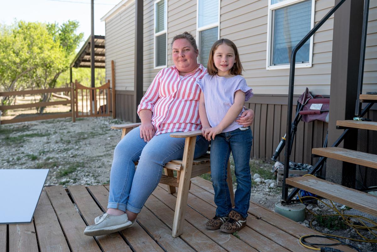 A woman sits in a chair with her arm around the waist of a child who stands next to her. They are outside a house.