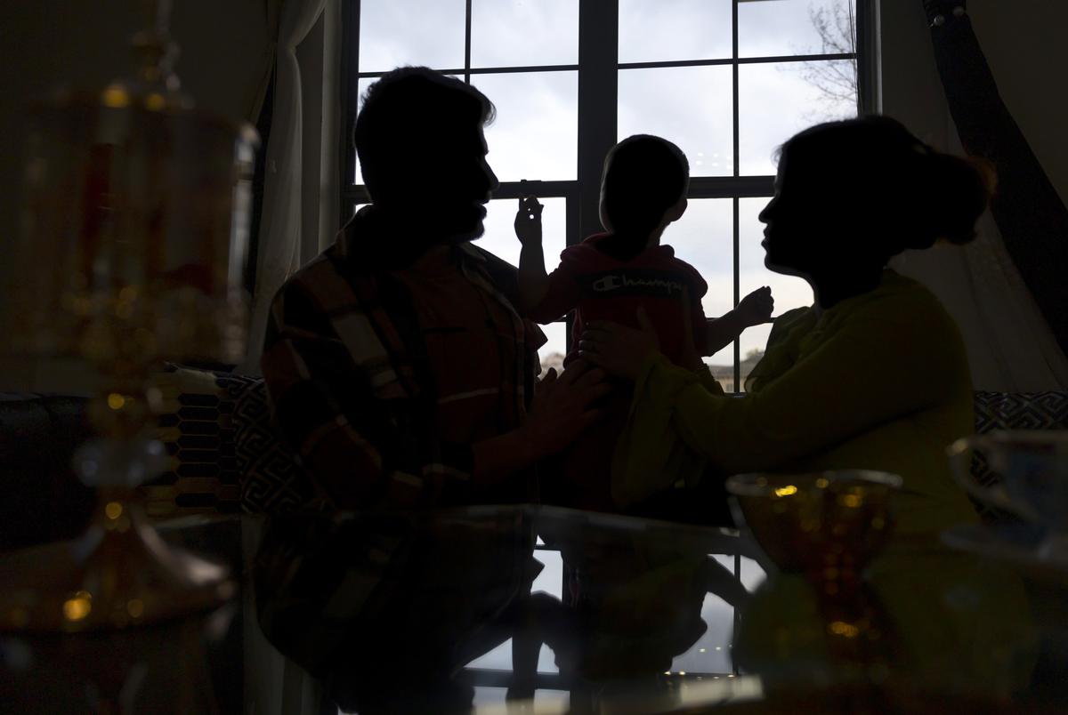 Two adults look at a child holding up a toy. All three are silhouetted against a sunny window and their faces cannot be seen.